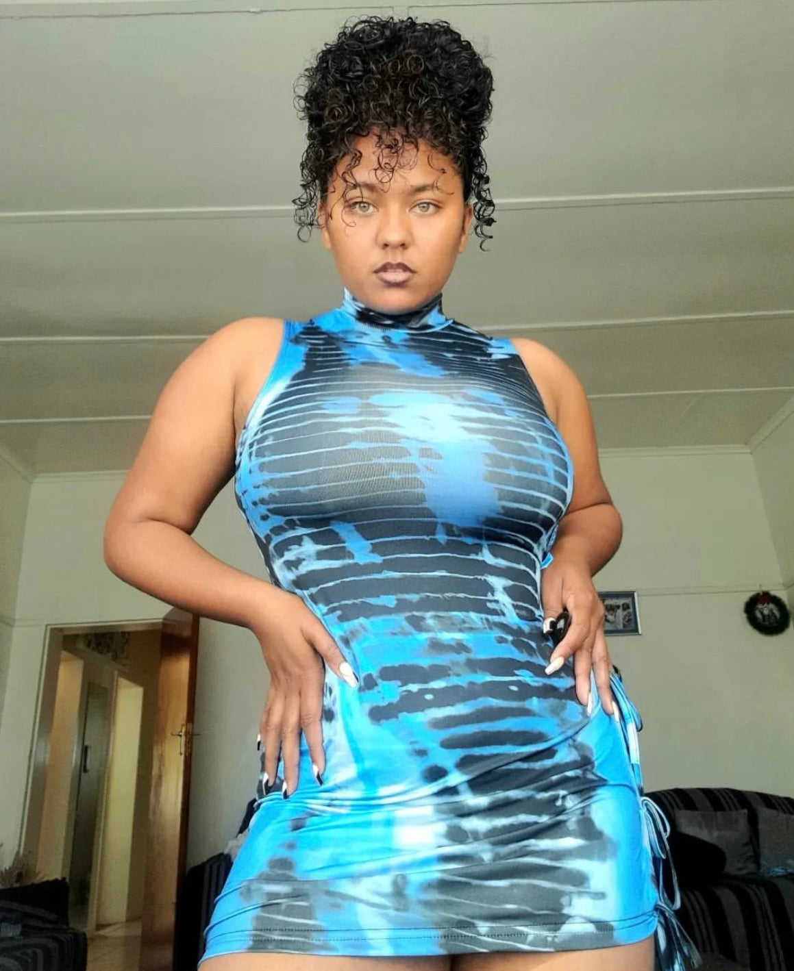 A women wearing a sleeveless, blue and black tie dye bodycon mini dress with side cut outs and drawstring details. The dress is form-fitting, showcasing the vibrant tie-dye pattern. 